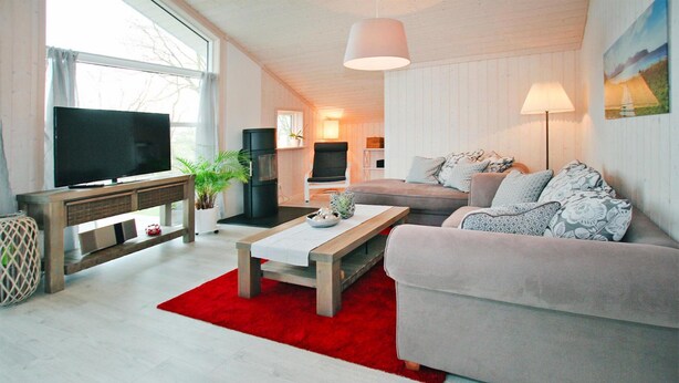 Ein modernes Wohnzimmer mit Fernseher, Couchtisch aus Holz, einem roten Teppich, einem grauen Sofa und einem großen Fenster.