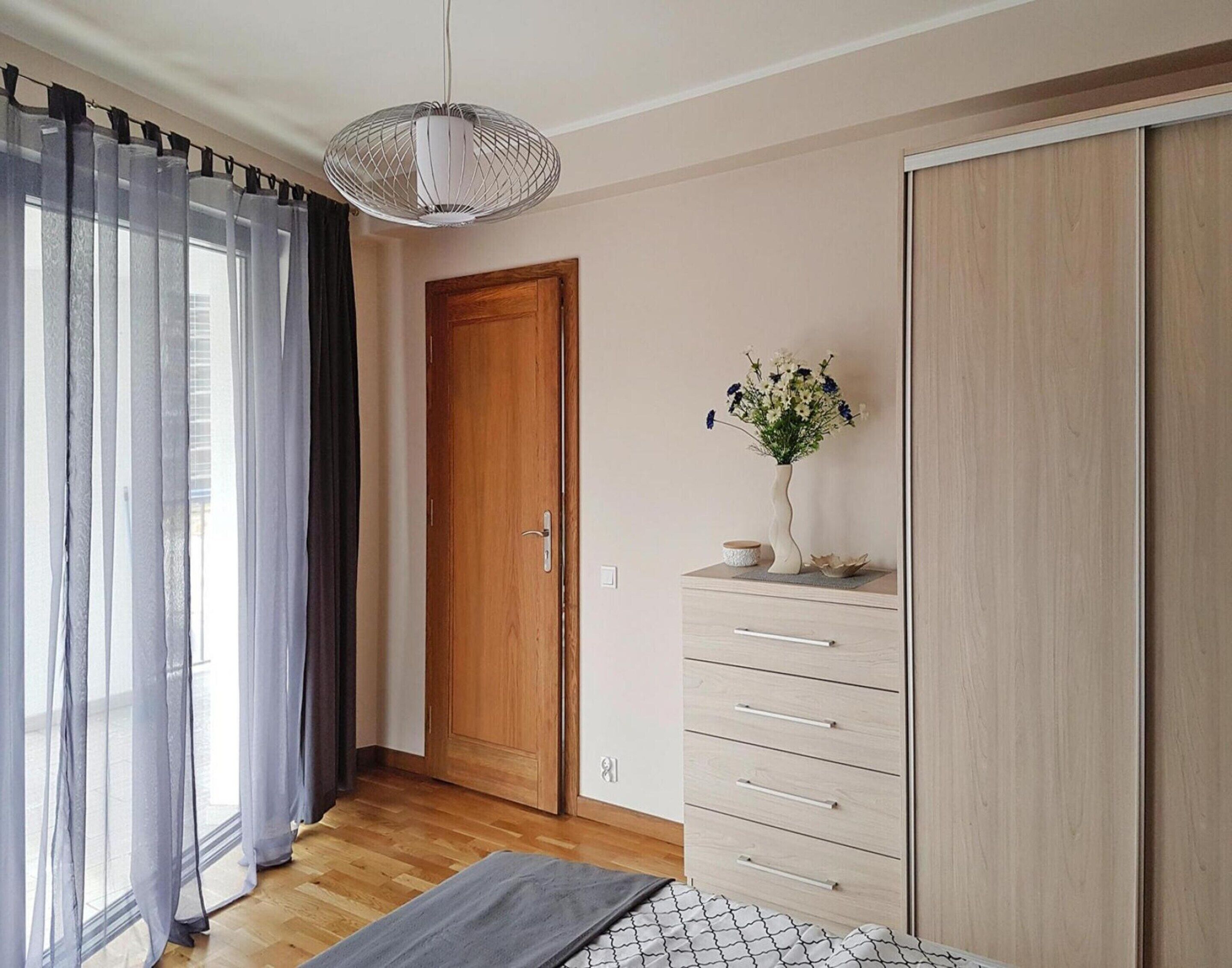 A bedroom with a wooden door, a dresser with drawers, a bedside table with a vase of flowers, and a hanging light fixture.