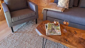 Una habitación acogedora con techo de madera, un sofá gris, un sillón gris y una pequeña mesa de centro de madera con dos copas de vino y una revista.