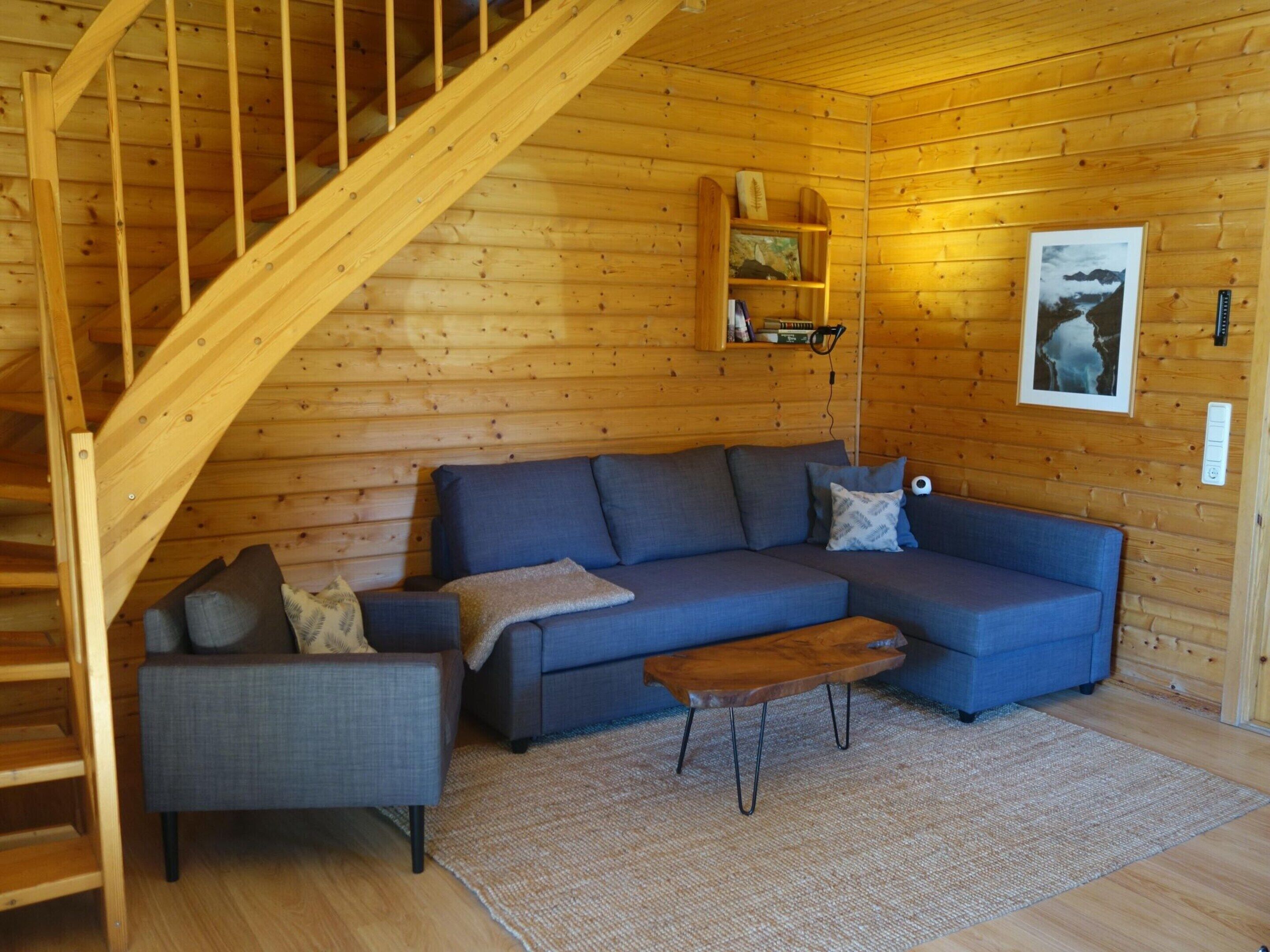 A wooden cabin interior with a staircase, a blue sofa set, a small table, and a mounted shelf with books and a picture.