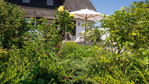 A thatched-roof building with a patio area featuring outdoor seating and a white umbrella. - Thatched Roof House in St.peter Ording (St. Peter-Ording)