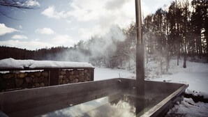Un bain à remous doté d’une structure en pierre et d’une haute cheminée d’où s’échappe de la vapeur, le tout entouré de neige et d’arbres.