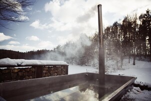 Un jacuzzi con estructura de piedra y una chimenea alta que emite vapor, rodeado de nieve y árboles.