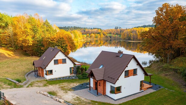 Deux maisons modernes avec des toits inclinés, une allée en gravier et un lac en arrière-plan.