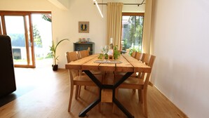 Un espace repas avec une table en bois, des chaises et une structure de support centrale noire. On aperçoit une plante en pot sur la gauche et une étagère avec des objets décoratifs à l’arrière-plan.