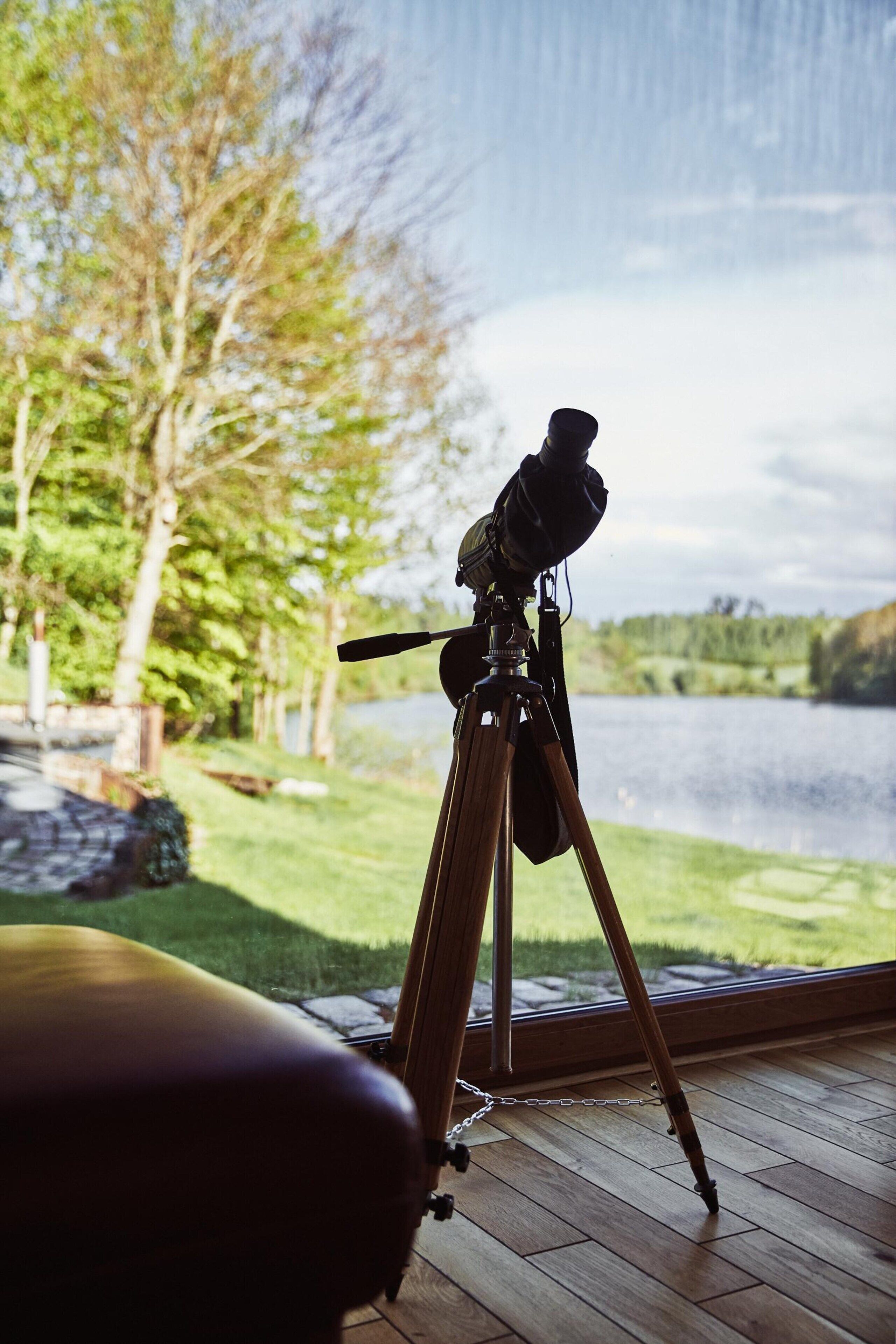 A telescope on a tripod inside a room with a view of a lake and trees outside.