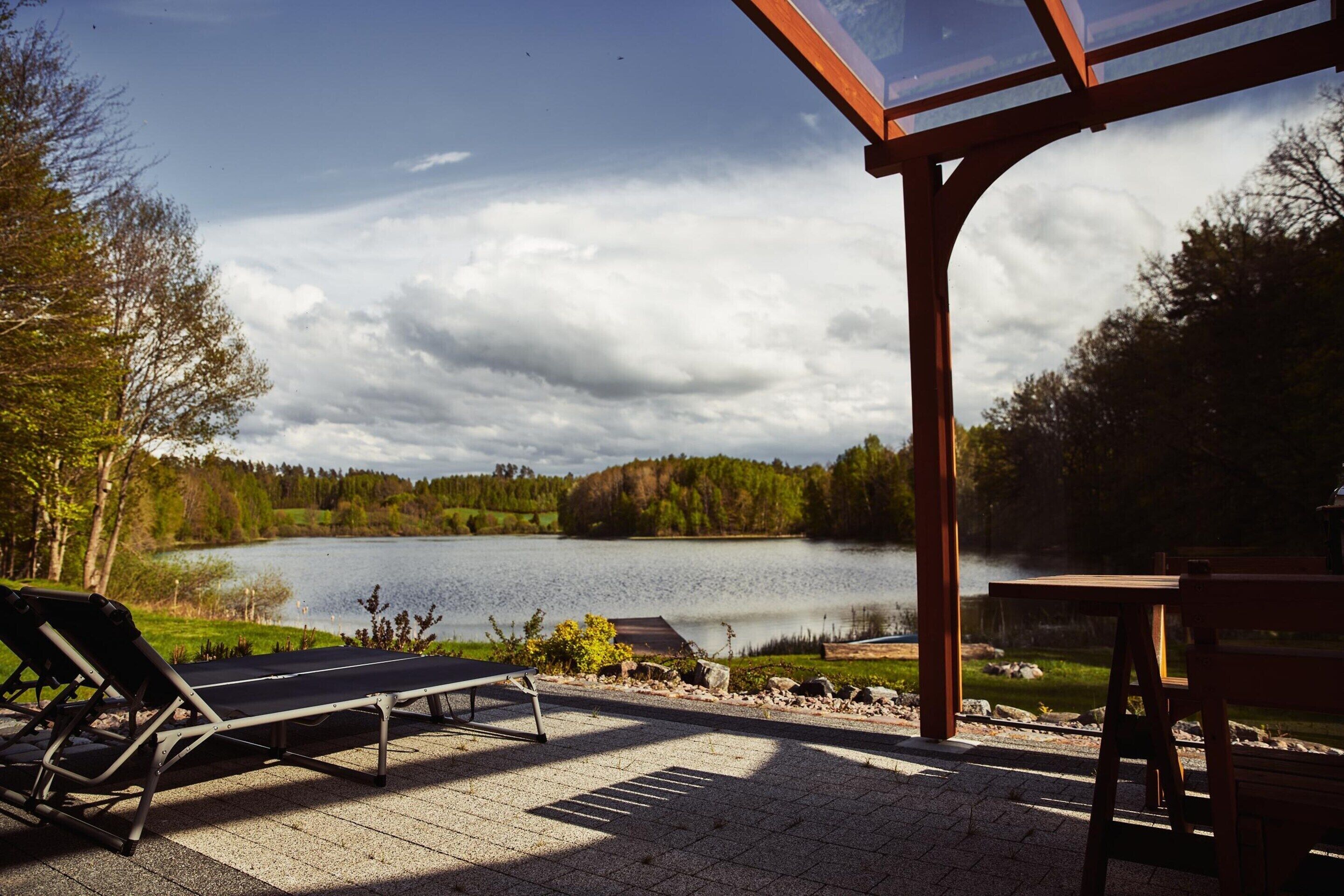 A lakeside patio with lounge chairs and a wooden table, overlooking a calm lake and forested hills.