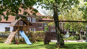 Une aire de jeux avec un toboggan, des balançoires et une structure d’escalade, entourée d’un champ d’herbe et d’arbres.