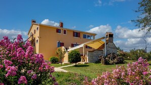 Una casa de dos pisos con una chimenea de piedra, un porche cubierto y un jardín con flores rosas.