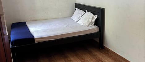 Una habitación pequeña con cama individual, suelo de madera, estructura de cama sencilla y pared lisa.