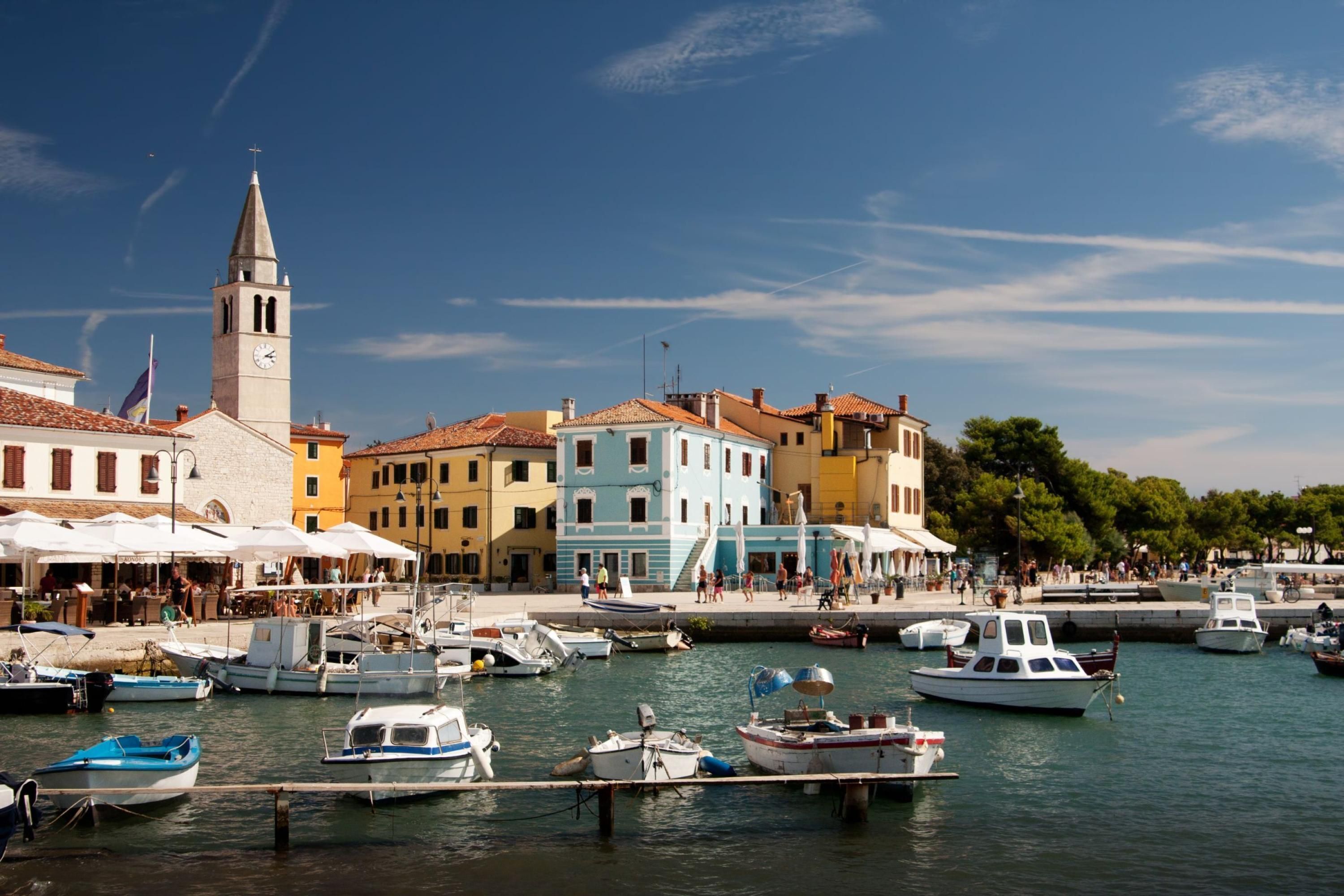 A marina with boats docked, a church with a bell tower, and colorful buildings along the waterfront.