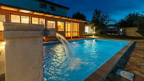 A swimming pool with a water feature, surrounded by a stone deck and a building with large windows and a tiled roof. - Pool Fun House Betiga 1 With gym (Vodnjan)
