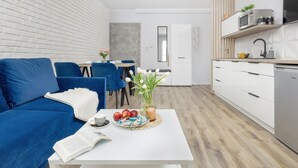 A modern living room with a blue sofa, a white coffee table with apples and a book, and a dining area with a table and chairs. - Comfortable Apartment, Pool, Close to the Beach (Rewal)