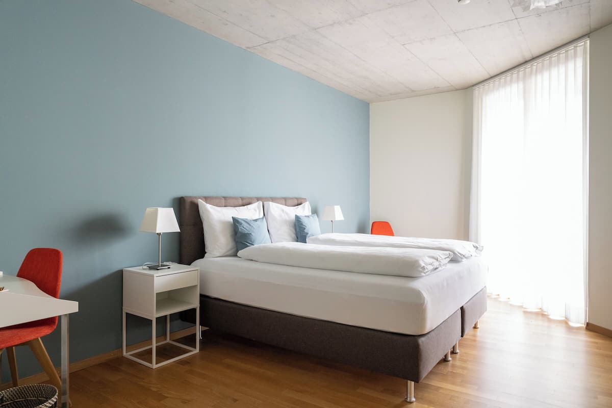 Un dormitorio con cama, mesita de noche, lámpara y una silla.