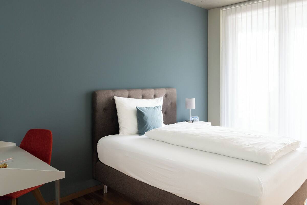 Una habitación de hotel con una cama, una mesita de noche con una lámpara, una silla roja y una mesa.