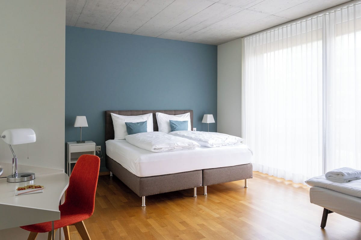 Habitación de hotel con cama, escritorio, silla, banco y un ventanal con persianas.