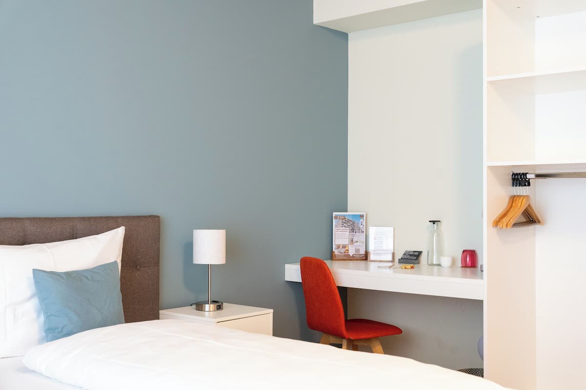 Una habitación de hotel con cama, escritorio, silla, mesita de noche, lámpara, un tablón de anuncios y perchero.