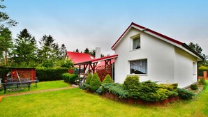 A two-story house with a red roof and white walls, surrounded by a well-maintained garden with various shrubs and a bench. - Spacious Cottage, Idyllic Right on the Lake, Spore (Szczecinek)