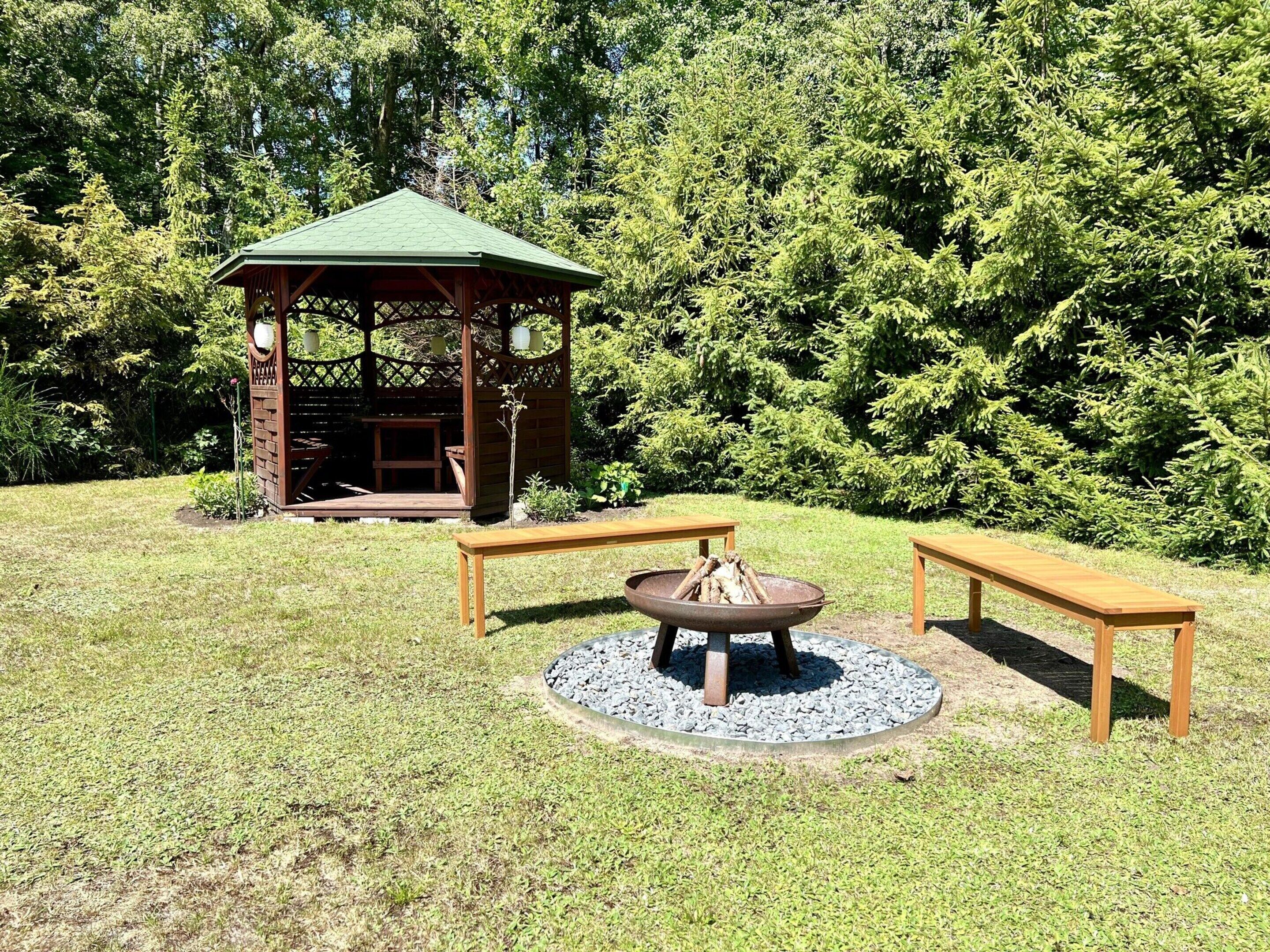 Ein Gazebo mit grünem Dach und Holzwänden, umgeben von einer Rasenfläche mit zwei Holzbanken und einer Feuerstelle in der Mitte.
