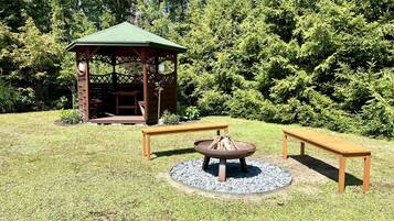 Ein Gazebo mit grünem Dach und Holzwänden, umgeben von einer Rasenfläche mit zwei Holzbanken und einer Feuerstelle in der Mitte.