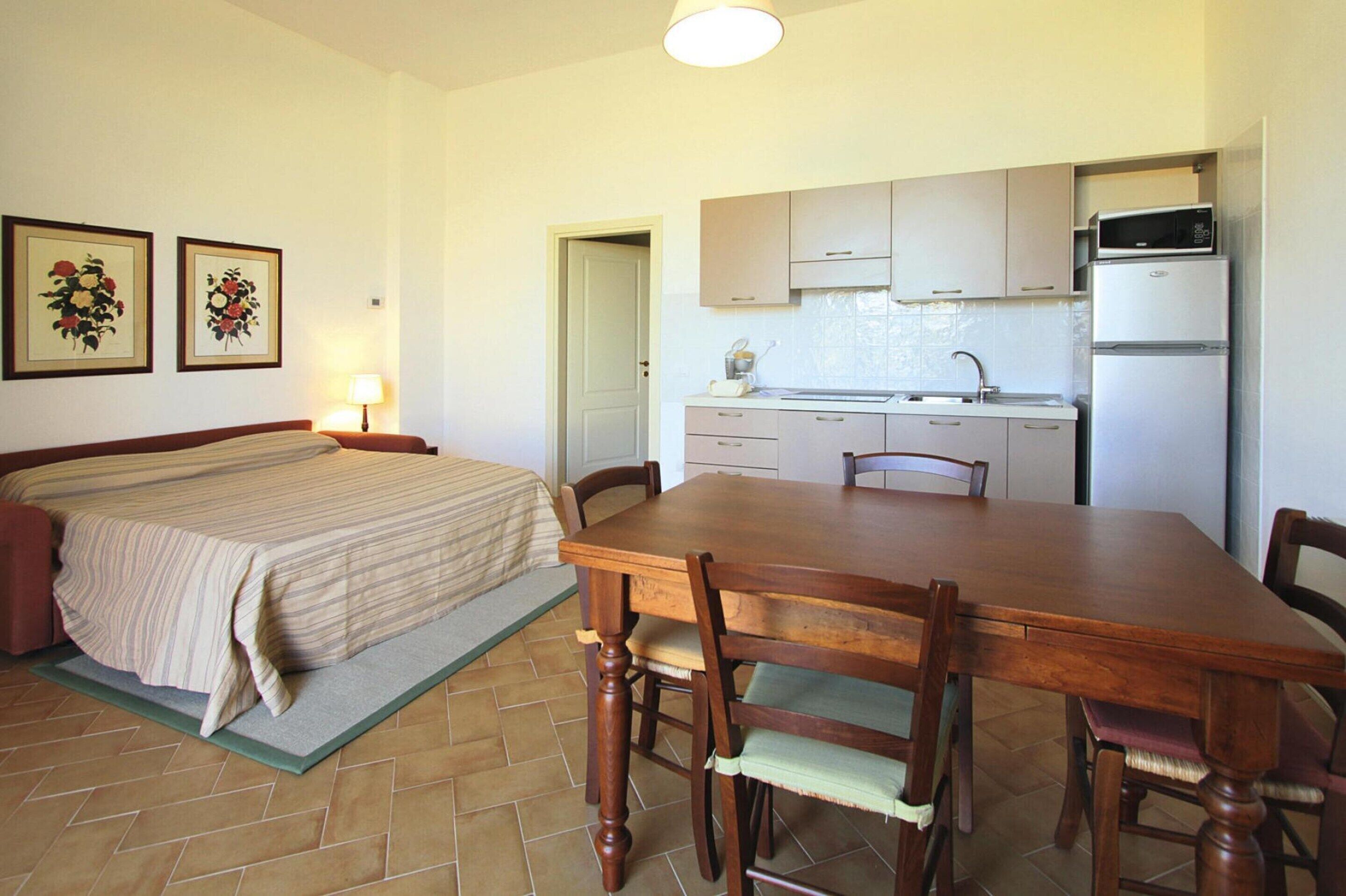 A bedroom with a bed, a wooden dining table with chairs, a kitchen with cabinets and appliances, and two framed pictures on the wall.