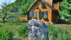 A wooden house with a balcony, surrounded by lush greenery and a large rock in the foreground.