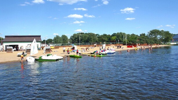 A lakeside scene with boats docked near the shore, a building with a green roof, and a sandy beach with people. - Holiday Appartment in Stepnica for 4 Persons (Stepnica)
