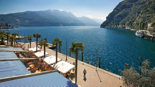 Ein Hotel am Wasserufer mit Außenbestuhlung, Palmen und Blick auf See und Berge.