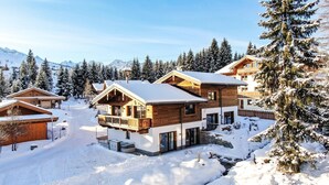 A chalet-style building with a wooden exterior and a balcony, surrounded by snow-covered trees and mountains in the background.