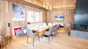 A dining area with a wooden table set for six, grey upholstered chairs, a wooden bench with cushions, a fireplace, and a television mounted on the wall.