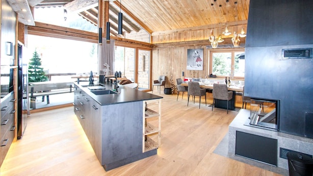 A modern kitchen with a large island, wooden ceiling beams, and a dining area with chairs and a table.
