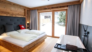 A modern bedroom with a wooden headboard, a large bed, a built-in bench, a bathtub, and a sliding glass door leading to a snowy outdoor area.