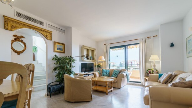 Ein geräumiges Wohnzimmer mit einer Couch, Sesseln und einem Couchtisch. Im Raum befindet sich ein Fernseher und eine Pflanze.