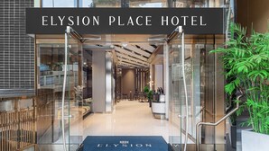 De ingang van Hotel Elysion Place met een modern ontwerp, met een glazen gevel, een receptiebalie en een blauw tapijt met de naam van het hotel.