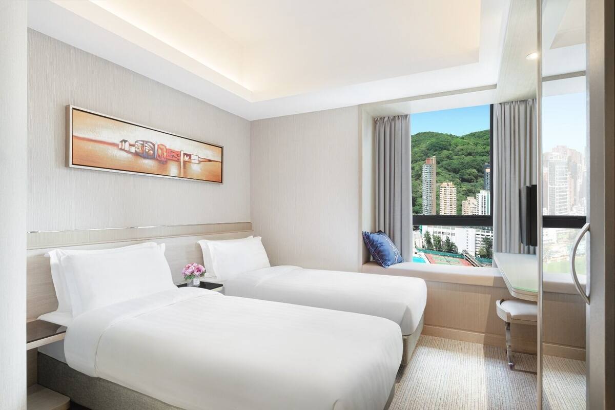 Une chambre d’hôtel avec deux lits, un bureau, une chaise, une télévision et une fenêtre donnant sur des bâtiments et des espaces verts.