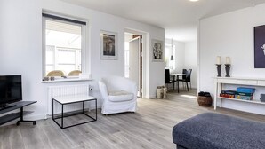 Ett modernt vardagsrum med en platt-TV, en vit fåtölj, en grå soffa, en matplats med bord och stolar samt en bokhylla med dekorativa föremål.