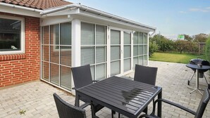 En täckt uteplats med ett bord och stolar, en grill och utsikt över ett hus med fasad av tegel och stora fönster.