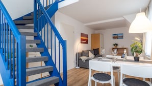 Ein moderner Wohnbereich mit einer blauen Treppe, einem Essbereich mit weißen Möbeln und einer gemütlichen Sitzecke mit Sofa und Kissen.