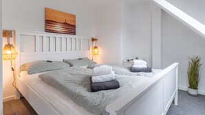 Ein modernes Schlafzimmer mit einem weißen Bett, zwei Nachttischlampen, einem Gemälde an der Wand und einer Topfpflanze.