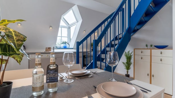 Ein Essbereich mit Tisch, gedeckt für zwei Personen, mit einer Wasserflasche, einer Flasche Schnirller und zwei Weingläsern. Im Hintergrund ist eine Treppe mit blauen Geländern zu sehen.