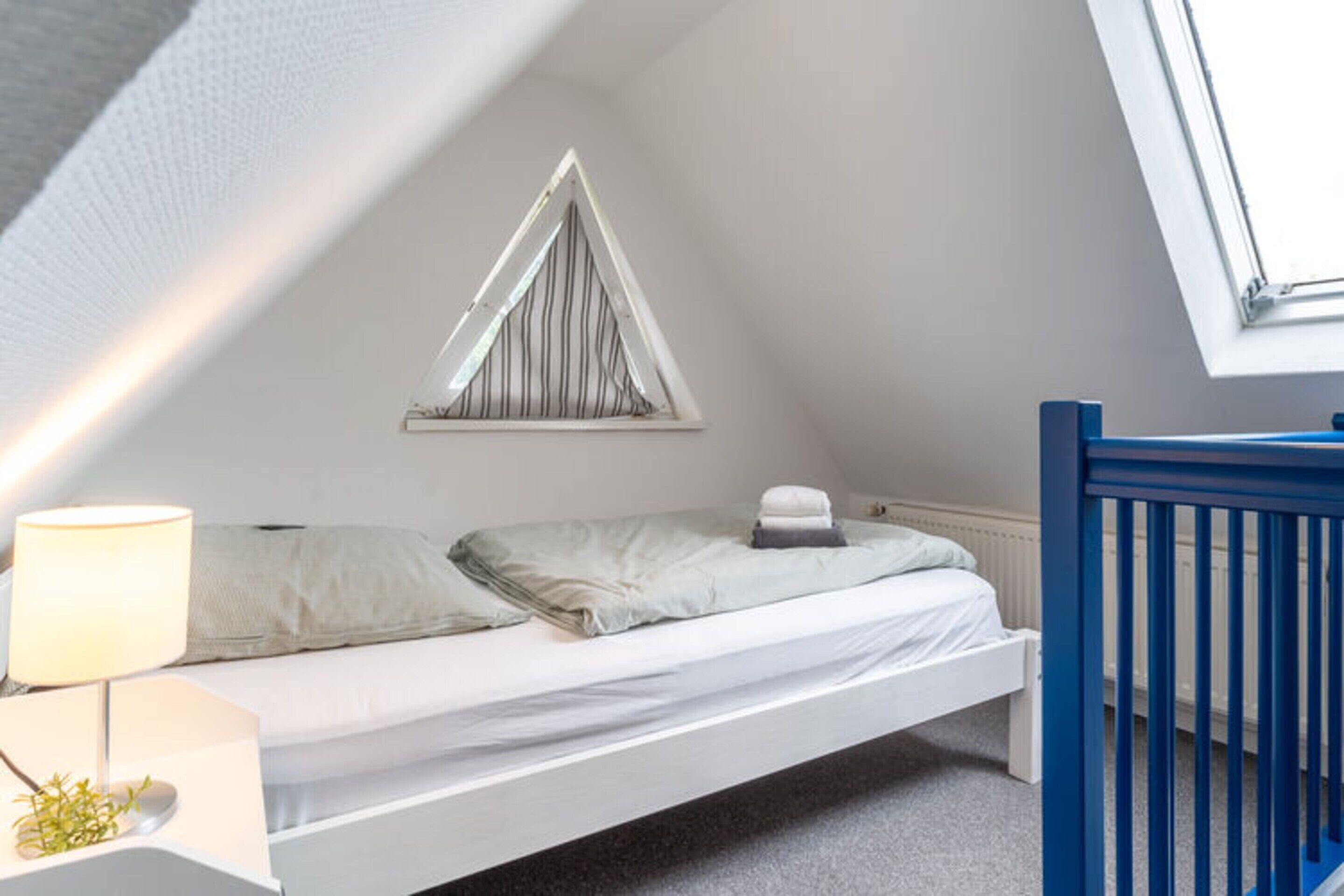 A small, neatly organized bedroom with a single bed, a window with a triangular view, a crib, and a bedside table with a lamp.