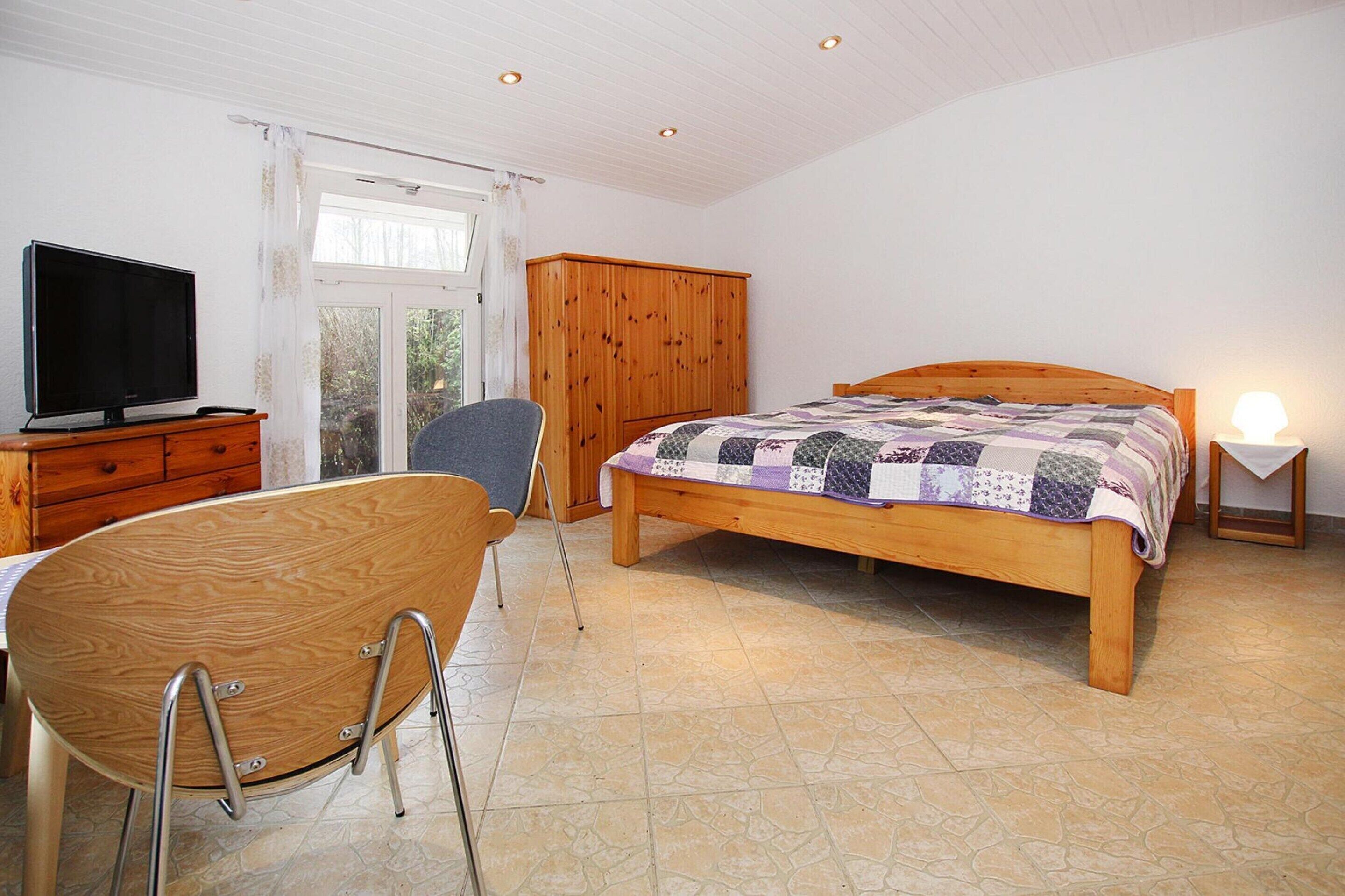 A bedroom with a wooden bed, a television on a wooden dresser, a chair, and a window with curtains.