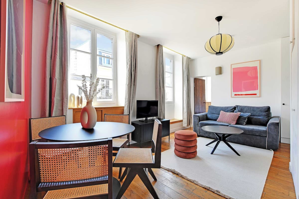 A modern living room with a round table, sofa, and ottoman. There is a television and a vase on the table.