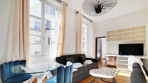 A modern living room with a glass table, blue chairs, a grey sofa, a TV, and a fireplace. - Résidence Paris BRETONNERIE (Paris)