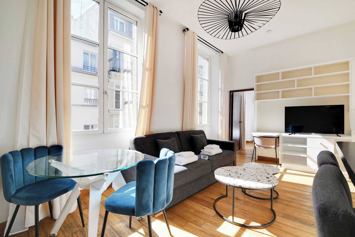 A modern living room with a glass table, blue chairs, a grey sofa, a TV, and a fireplace.
