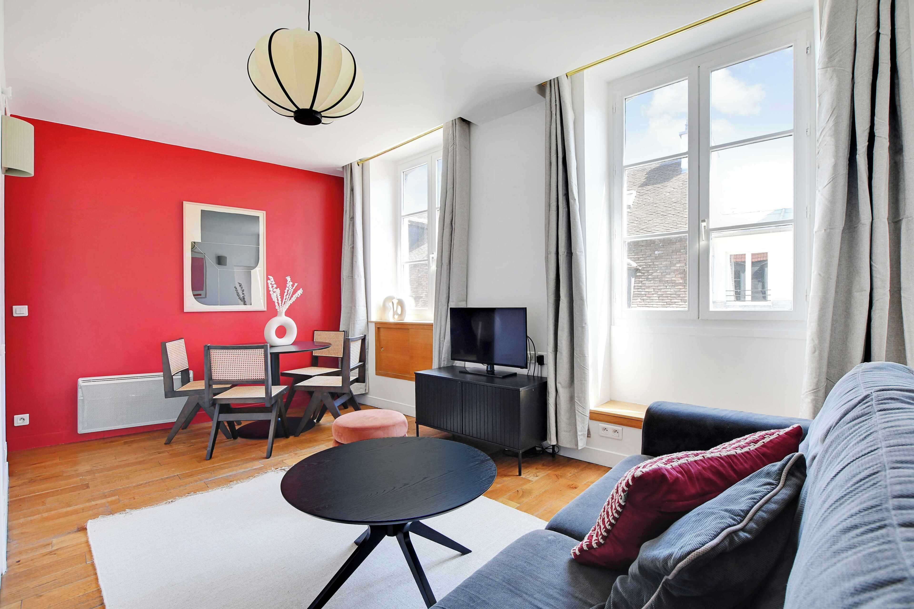 Una sala de estar con una pared roja, un sofá gris, una mesa de centro negra y un televisor sobre un mueble negro.
