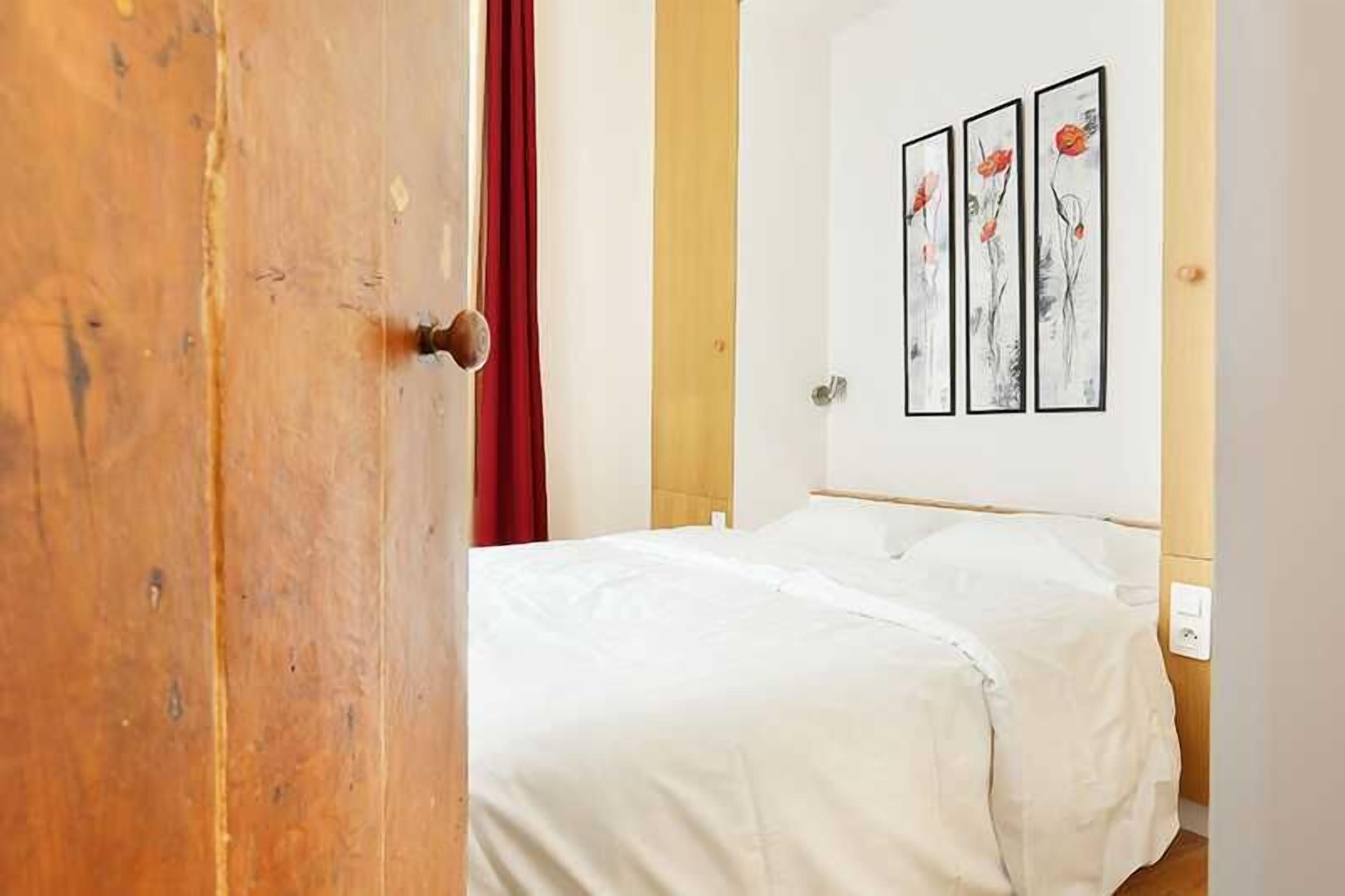 A hotel room with a wooden door, a bed with white bedding, and three framed artworks on the wall.