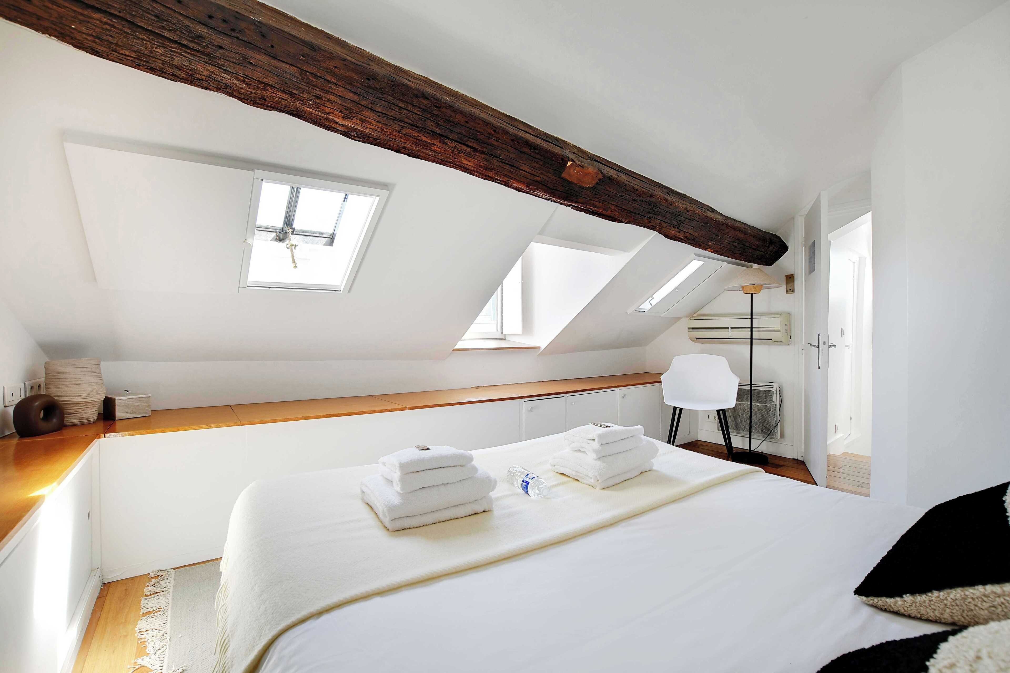 A bedroom with a sloped ceiling, skylights, a bed with white linens, a bedside table, a lamp, and a chair.