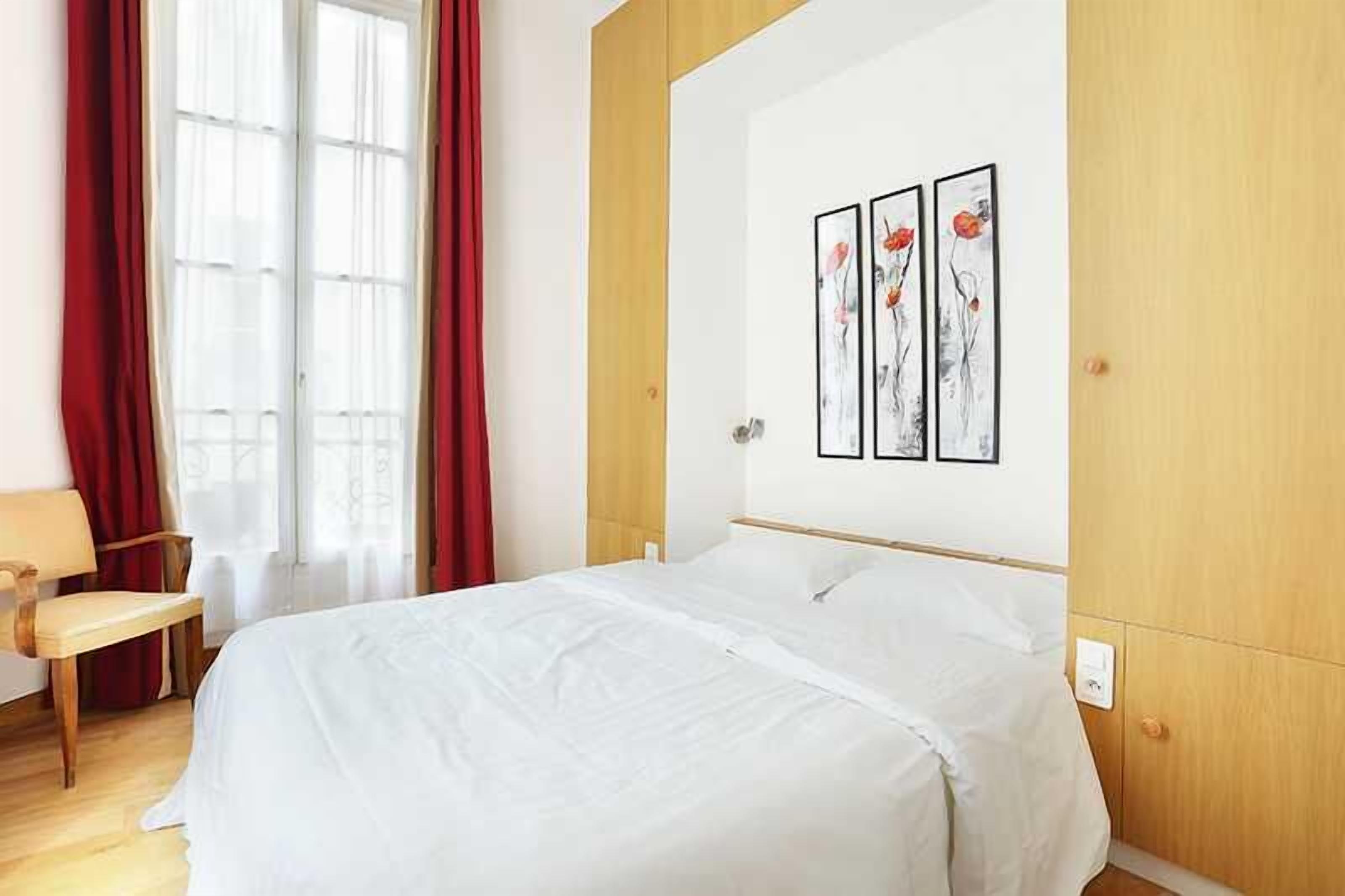 Una habitación de hotel con una cama, una silla, un ventanal con cortinas rojas y tres cuadros en la pared.