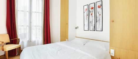 Una habitación de hotel con una cama, una silla, un ventanal con cortinas rojas y tres cuadros en la pared.